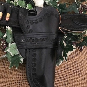 May include: A black leather holster with intricate stitching and a decorative design. A silver revolver with a white handle is in the holster. The holster is attached to a brown leather belt with a silver buckle.