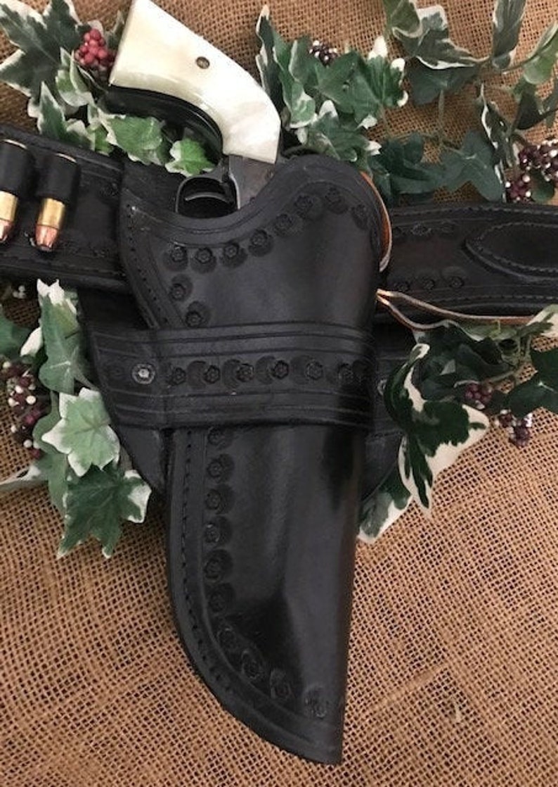 May include: A black leather holster with intricate stitching and a decorative design. A silver revolver with a white handle is in the holster. The holster is attached to a brown leather belt with a silver buckle.