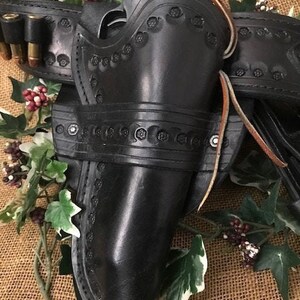 May include: A black leather holster with intricate tooling and a silver buckle. The holster is designed to hold a pistol and has a belt loop for attaching to a belt.