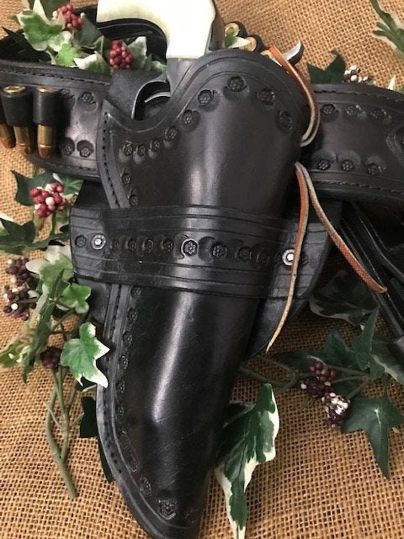 May include: A black leather holster with intricate tooling and a silver buckle. The holster is designed to hold a pistol and has a belt loop for attaching to a belt.