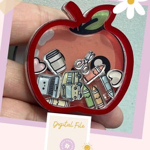 May include: A red apple-shaped shaker charm filled with school-themed illustrations, including a laptop, crayons, and scissors. The charm has a clear center and the words "Digital File" are displayed on a small card.