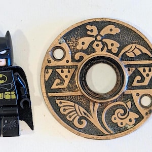 May include: Ornate, circular bronze door hardware with a central hole and decorative floral and geometric patterns. The hardware is next to a Batman Lego figurine.