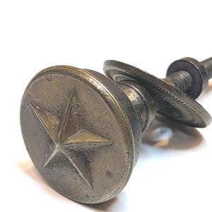 May include: A round, antique brass-toned metal knob featuring a raised five-pointed star design on its face. The hardware includes a threaded bolt, a circular washer, and a nut attached to the back, showing a weathered, metallic finish throughout.
