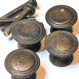 May include: A set of three round cabinet knobs with a weathered, dark brass-toned finish and a raised circular border detail. The knobs feature a tapered base and are accompanied by three matching metal mounting screws with threaded shafts.