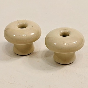 May include: Two off-white ceramic insulators with a hole in the center. The insulators are shaped like a mushroom with a rounded top and a short stem.