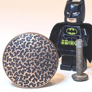 May include: A circular knob with a textured, copper-colored finish featuring a black, organic, maze-like pattern. Next to it stands a black Batman minifigure and a small, threaded metal bolt, providing a sense of scale for the hardware piece.