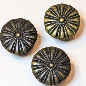 May include: Three round metal buttons with a dark brass finish. Each button features a raised center dot surrounded by radiating grooves that extend to the outer edge, creating a sunburst pattern across the surface of the circular fasteners.