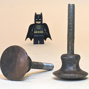 May include: Two dark brown metal furniture levelers with threaded stems. One leveler is lying on its side, while the other stands upright on its base. A small black Batman figurine stands in the background between the two metal components.
