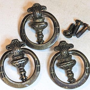 May include: Three circular metal drawer pulls with a tarnished brass finish, each featuring a central decorative post and a swinging ring handle. The set includes three matching metal screws with flat heads for installation purposes.