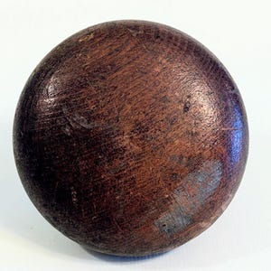 May include: A round, wooden ball with a dark brown finish. The surface shows visible wood grain and some areas of discoloration. The ball appears to be smooth and well-worn, suggesting it has been handled.