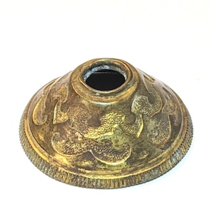 May include: A small, circular, brass-colored metal component with a conical shape and a central hole. The surface features an embossed, swirling pattern and a textured, ridged edge. A partial black plastic figurine is visible in the lower left corner.