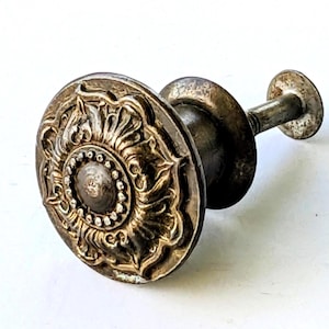 May include: A round metal cabinet knob with a tarnished silver finish featuring an embossed floral pattern on the face, a central circular stud surrounded by a beaded border, and a threaded metal post extending from the back for mounting purposes.