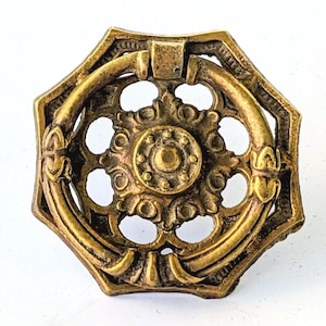 May include: A brass-toned metal drawer pull featuring an octagonal backplate with an intricate openwork design and a central circular medallion. A hinged circular ring handle is attached to the front, displaying a weathered, aged finish.
