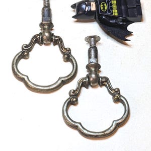 May include: Two silver-toned metal drawer pulls with ornate, scalloped ring handles and a small Batman minifigure with a black cape, yellow utility belt, and a black cowl, positioned next to the hardware for a sense of scale.