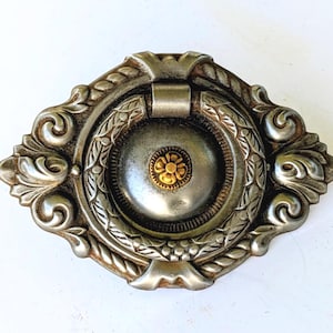 May include: An oval-shaped metal drawer pull featuring a silver-toned finish with ornate scrollwork and rope-like detailing. A central circular ring hangs over a domed silver center, which is accented by a small, raised gold-colored flower motif.