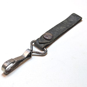 May include: A dark gray leather key fob strap featuring a weathered texture and a single metallic snap button. The strap is attached to a silver-toned metal swivel lobster clasp hook with a triangular connector, showing signs of wear and age.