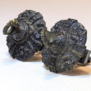 May include: Two dark, weathered metal drawer knobs with a circular, textured floral pattern and a small, protruding loop at the center. Each knob features a tarnished, aged finish with visible relief details and a threaded metal bolt on the back.