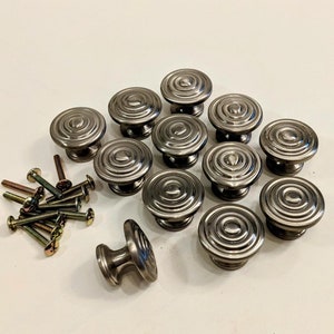 May include: A set of 12 silver-toned round cabinet knobs with a concentric circle design. The knobs are arranged in a circle with a single knob in the center. The knobs come with screws.