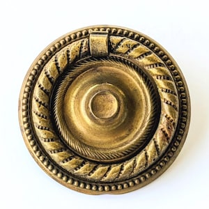 May include: A circular brass drawer pull featuring a central recessed disc surrounded by a textured rope-style ring. The outer edge of the hardware is decorated with a beaded border, and the entire piece has a weathered, antique gold finish.