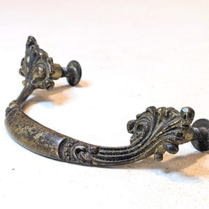 May include: A curved metal drawer pull with an aged bronze finish. The handle features ornate, leaf-like scrollwork at both ends and two circular mounting posts with screws extending from the back to attach to a cabinet or furniture surface.