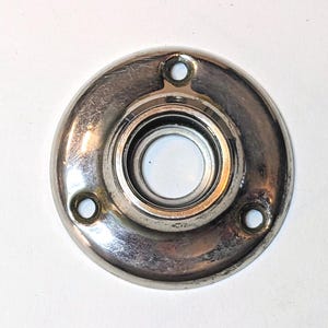 May include: A circular, polished chrome-finished metal door rosette featuring a central opening for a spindle and three evenly spaced mounting holes along the outer edge. The hardware has a reflective, metallic surface with visible signs of wear.