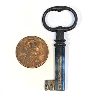 May include: A dark, weathered metal skeleton key with an oval bow and a cylindrical shaft, positioned next to a copper-colored United States one-cent coin for scale. The key features a simple bit at the bottom and a distinct collar below the bow.