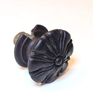 May include: A dark brown, circular cabinet knob featuring a raised, fluted, flower-like pattern radiating from a central point. The knob has a thick, rounded edge and a textured, metallic-looking base that extends outward from the back.