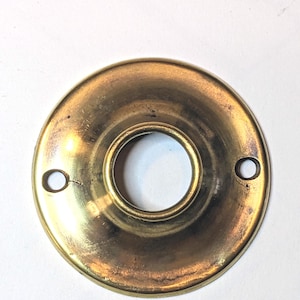 May include: A circular brass door knob rosette with a polished gold-toned finish. The hardware features a central circular opening for a spindle and two smaller, opposing holes on the outer edge for mounting screws, showing a smooth surface.