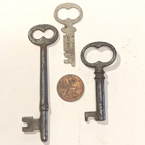 May include: Three antique metal keys with oval bows and distinct bit patterns arranged around a copper-colored United States one-cent coin. The keys exhibit a weathered, dark grey patina, showing signs of age and oxidation on their surfaces.