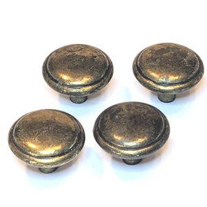 May include: A set of four round cabinet knobs with a distressed antique brass finish. Each knob features a smooth, domed top surface with a raised circular border and a central mounting post, displaying a weathered, dark metallic texture.