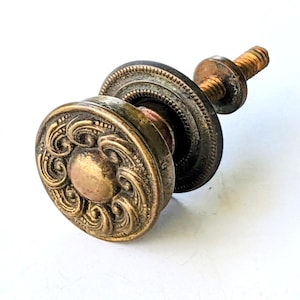 May include: A round brass cabinet knob featuring an embossed swirling scroll pattern on the face. The hardware includes a circular base plate and a threaded metal bolt extending from the back for installation, showing a weathered metallic finish.