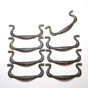 May include: A set of nine dark, weathered metal drawer pulls arranged in two columns. Each pull features a curved, horizontal handle with distinct, upward-curving ends designed for mounting. The metal surface shows a mottled, aged patina.