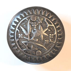 May include: A round, dark metallic button featuring an embossed floral and leaf pattern in the center. The outer edge is decorated with a series of small, radiating triangular notches. The surface has a weathered, aged finish with dark gray tones.