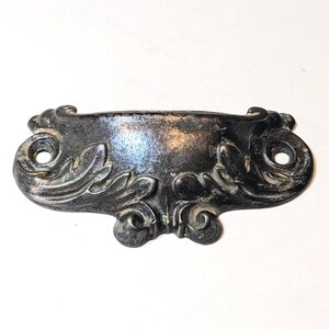 May include: A dark metallic drawer pull with a weathered finish, featuring a central cup-shaped handle and decorative scrollwork on both sides. The piece includes two circular screw holes on the outer edges for mounting to a furniture surface.
