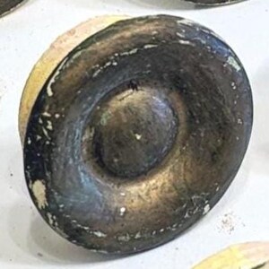 May include: A circular cabinet knob with a distressed finish featuring a dark, weathered bronze-toned center and a lighter, cream-colored outer edge. The surface shows signs of wear with visible paint chipping and a textured, aged appearance.