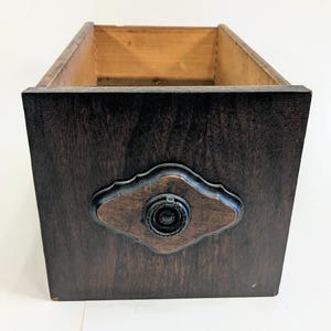 May include: A dark brown wooden drawer with a decorative metal pull. The drawer has a rectangular shape with a lighter wood interior. The metal pull is a dark color with a decorative design.