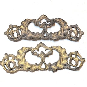 May include: Two matching metal drawer pulls with an aged gold finish and a dark patina. Each piece features an intricate, symmetrical openwork design with leaf-like motifs and circular mounting holes on both ends for attachment to furniture.