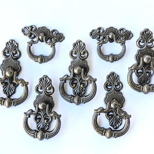 May include: A collection of seven bronze-toned metal drawer pulls featuring ornate, symmetrical scrollwork designs. Each piece consists of a decorative backplate with an attached swinging ring handle, showcasing a dark, aged metallic finish.