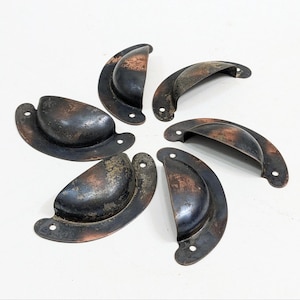 May include: Six antique, half-moon shaped drawer pulls. The metal pulls have a dark, aged appearance with areas of rust-colored patina. Each pull has two screw holes for mounting. The pulls are arranged in a circular pattern.