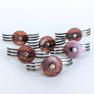 May include: A collection of five metal drawer pulls, each featuring a circular, marbled reddish-brown resin inlay centered with a small metallic stud. The base of each pull consists of three parallel, curved metal arms extending outward.