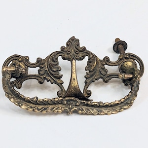 May include: Ornate antique brass drawer pull with a decorative, scrolling design. The pull features a central vertical element and a curved handle. Includes a screw for mounting. Ideal for vintage furniture restoration.