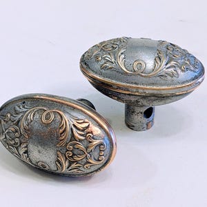 May include: Two antique oval door knobs with ornate floral designs. The knobs are silver with gold-colored accents and show signs of wear. One knob is attached to a stem. The door hardware is likely made of metal.