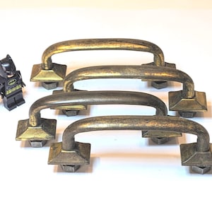 May include: Four arch-shaped cabinet handles with a distressed antique brass finish. Each handle features a curved bar attached to two square bases with hexagonal nuts underneath. A small black Batman figurine stands to the left for scale.
