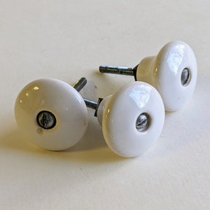 May include: Three white ceramic cabinet knobs with silver screws. The knobs are round and have a slight indentation in the center.
