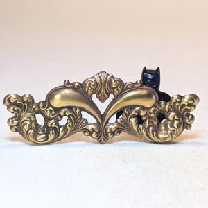 May include: A brass-colored metal drawer pull featuring an ornate, symmetrical scrollwork design with two smooth, curved central elements. A small, black, plastic figurine resembling a masked superhero is positioned behind the right side.