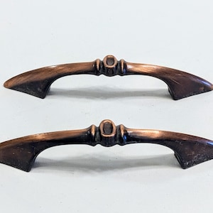 May include: Two antique-style cabinet pulls with a copper finish. The pulls have a curved shape with a decorative design in the center.
