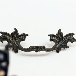 May include: Ornate, antique-style drawer pull with a dark bronze finish. The decorative handle features a scrolling leaf design, perfect for adding a vintage touch to furniture. The pull measures approximately 6 inches in length.