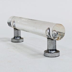 May include: A clear, cylindrical cabinet pull with a transparent body and two silver-colored metal posts. The hardware is designed for easy installation on drawers or cabinets. The overall design is modern and minimalist.