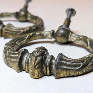 May include: Two antique-style brass drawer pulls featuring a circular ring design with ornate, textured detailing. Each pull includes a central mounting post and a threaded screw extending from the back, showcasing a weathered, aged metal finish.
