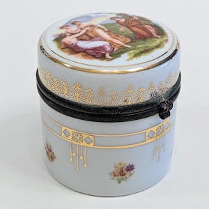 May include: A white porcelain trinket box with a gold trim and a lid featuring a painted scene of three women in a landscape. The box has a hinged lid with a metal clasp.